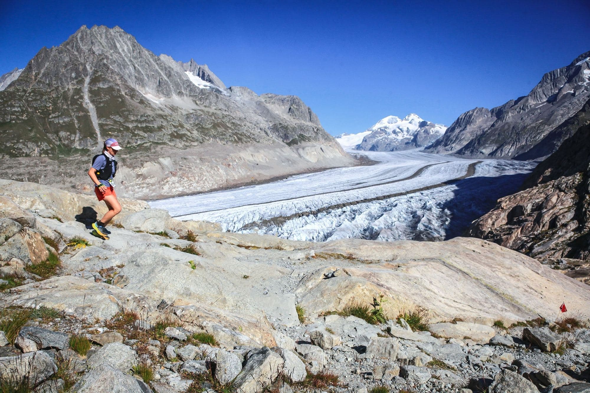 A trail runner on a rocky path above a long glacier, snowy peaks behind