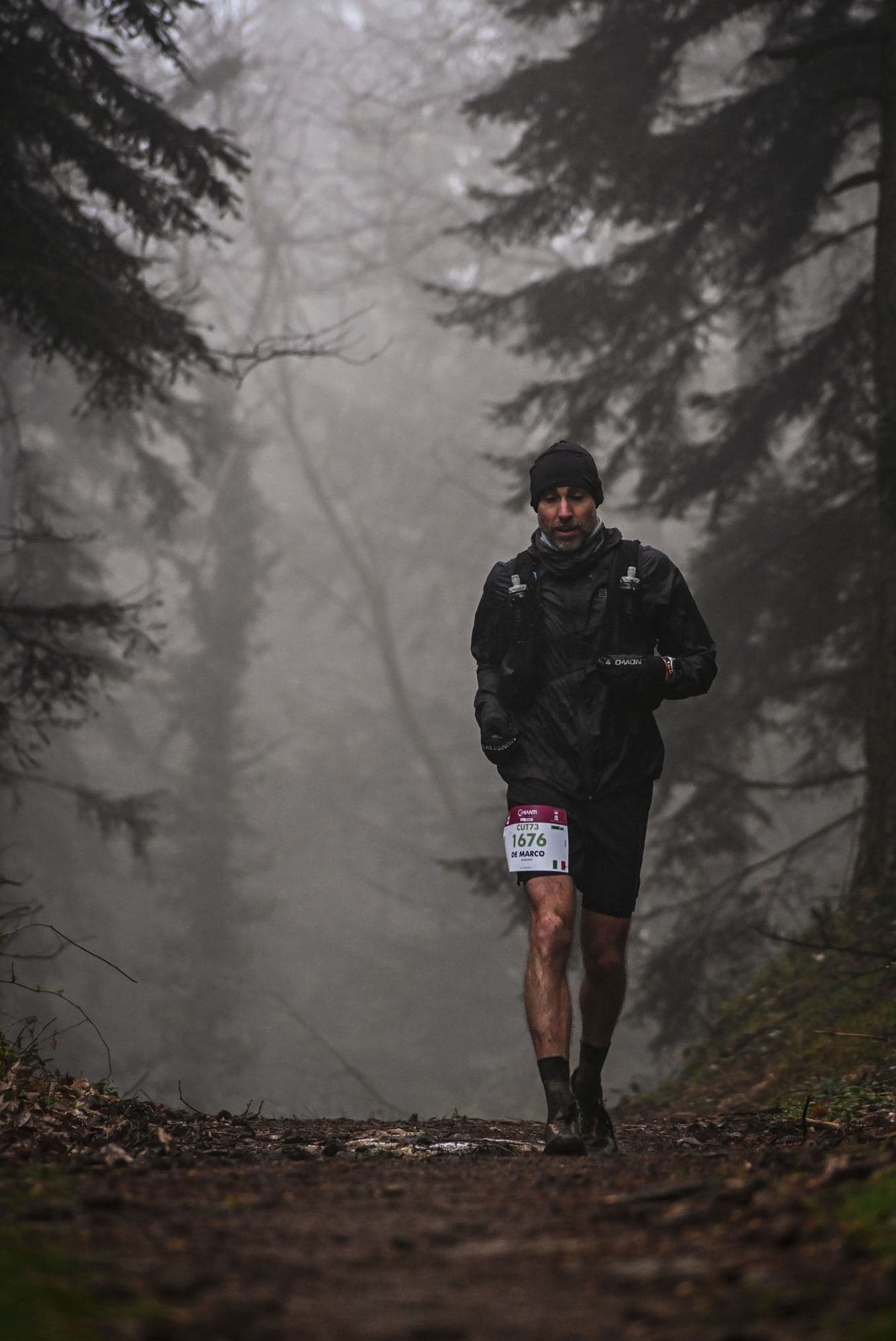 Trail runner con il pettorale che scende da un sentiero nel bosco, nella nebbia